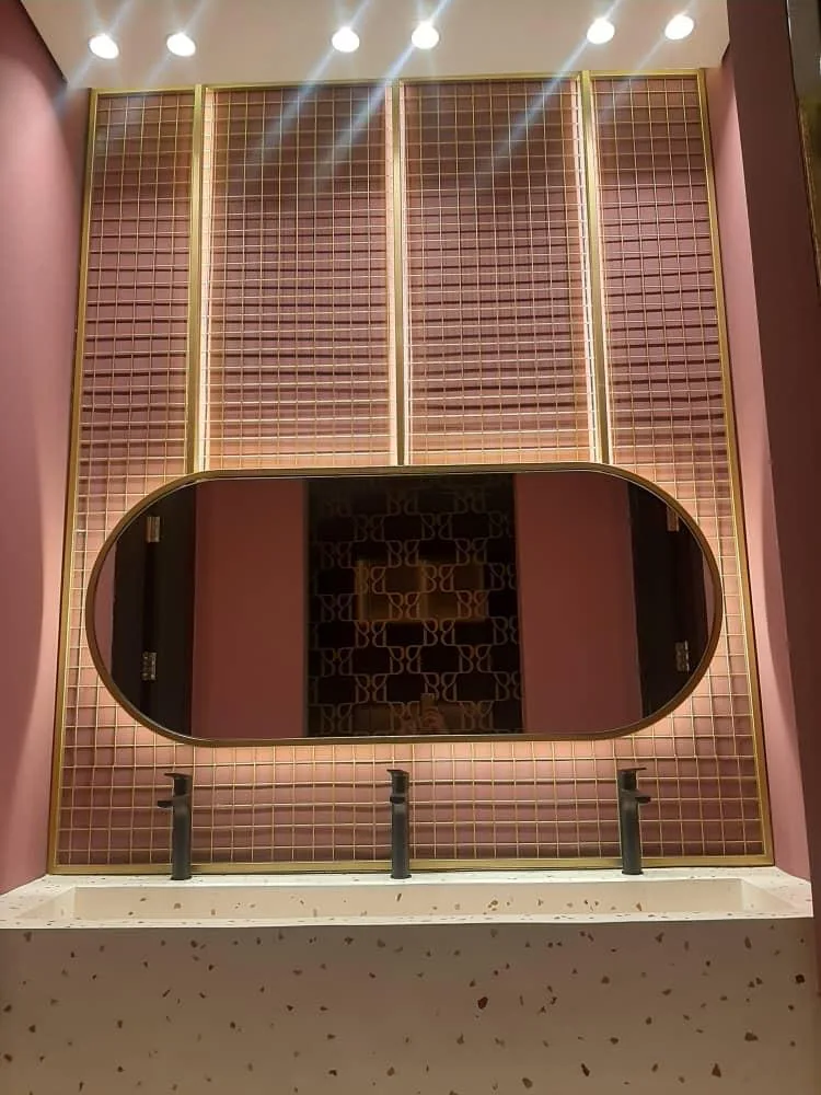 Washroom — oval brass-framed mirror on a pink tiled wall above a terrazzo trough basin